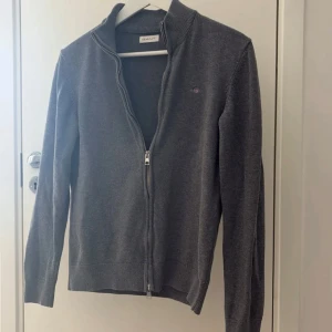 Grå kofta från GANT med dragkedja - STRL-Xs passar någon runt 165-170      Mörkgrå kofta från GANT med hel dragkedja och liten broderad logga på bröstet. Tröjan är ribbad vid krage, ärmslut och nederkant. Perfekt för lager-på-lager och har en clean, klassisk look.      Nypris 1200kr. 