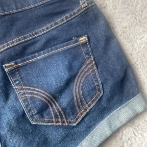 Mörkblå jeansshorts med uppvik - Snygga mörkblå jeansshorts med klassiska kontrastsömmar och uppvikta benslut. Shortsen har fem fickor och bälteshällor, perfekt för en chill sommarlook. Bakfickorna har dekorativa sömmar som ger extra stilpoäng.