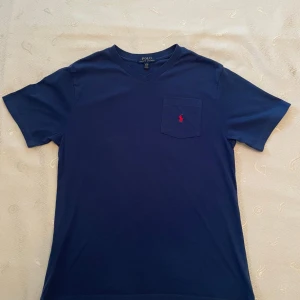 Blå t-shirt från Polo Ralph Lauren - Mörkblå t-shirt från Polo Ralph Lauren med v-ringning och bröstficka. Klassisk röd broderad logga på fickan. Skön passform och tillverkad i mjuk bomull. Perfekt för en clean och stilren look. 14-16 år men passar även S