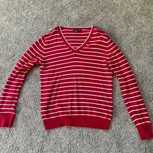 Randig röd tröja från Isolde - Snygg röd tröja från Isolde med vita horisontella ränder och v-ringad hals. Tröjan har långa ärmar och ribbade muddar vid ärmslut och nederkant. Perfekt för dig som gillar klassisk stil med en färgklick.