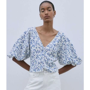 Blommig blus med puffärm och v-ringning - Superfin blus i vitt med blått blommönster, v-ringad framtill och knappar längs hela framsidan. Puffiga, korta ärmar ger en trendig siluett. Perfekt till ljusa jeans eller kjol för en fräsch look.