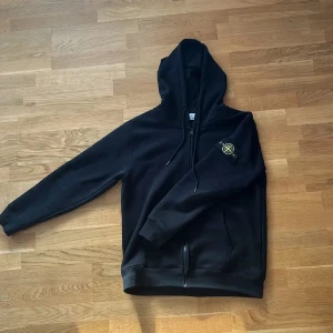 Svart hoodie från Stone Island - Svart hoodie från Stone Island med dragkedja framtill och klassisk logga på vänster ärm. Tröjan har huva med snörning och ribbade muddar. Perfekt för dig som gillar streetwear och vill ha en stilren look.