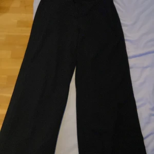 Svarta vida kostymbyxor - Snygga svarta kostymbyxor med vida ben och klassisk passform. Byxorna har hög midja, bälteshällor och stängs med knapp och dragkedja. Perfekta för en stilren look och passar till många olika outfits.