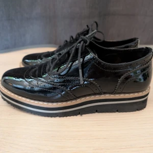Svarta lackloafers med platåsula - Snygga svarta loafers i lack med klassiska broguedetaljer och snörning. Skorna har en markerad platåsula med kontrastfärgade ränder och rund tå. Perfekta för dig som vill sticka ut med en dressad men edgy look.