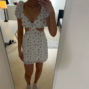 Vit blommig kortklänning  - Vit kortklänning med blått blommmönster Klänningen har puffärmar, cut-outs i midja.