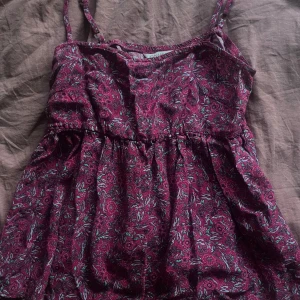 Blommig vinröd linnetopp från ONLY - Supersöt vinröd linnetopp från ONLY med blommigt mönster i grönt och rosa. Tunt och luftigt material, smala axelband och lätt rynkad midja för en avslappnad look. Perfekt för varma dagar när du vill känna dig både chill och snygg.