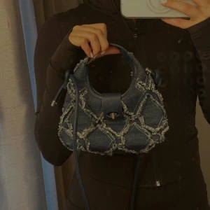 Jeans handväska med fransiga detaljer - Cool handväska i jeansmaterial med fransiga, rutiga detaljer och mörkblå läderremmar. Väskan har ett snyggt spänne framtill och både handtag och axelrem, perfekt för en trendig look.