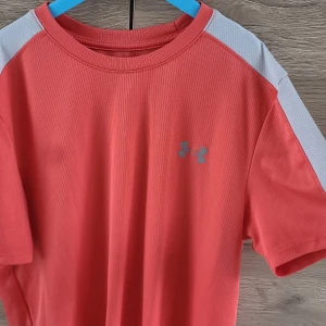 Under Armour tränings-tshirt - Sportig t-shirt från Under Armour med grå logga på bröstet och vita paneler på axlarna. Tillverkad i tekniskt, lätt och ventilerande material med diskret ribbad struktur. Perfekt för dig som gillar att träna och vill ha en snygg look.