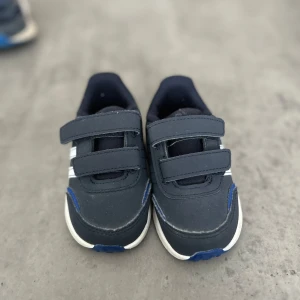 Mörkblå adidas sneakers med kardborre - Snygga mörkblå sneakers från adidas med klassiska vita ränder på sidorna och blå detaljer. Skorna har praktiska kardborreband och vit sula. Perfekta för en sportig och avslappnad stil.