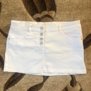 Vit minikjol - Säljer en snygg vit kort jeanskjol med fyra silvriga knappar framtill och klassiska fickor både fram och bak. 