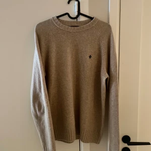 Beige stickad tröja från Morris - Snygg beige stickad tröja från Morris med rund halsringning och svart broderad logga på bröstet. Tröjan har ribbade muddar vid ärmslut och nederkant. Materialet är 100% lamull vilket är ett väldigt exklusivt och bekvämt material. Storlek S passar S/M nyskick! Endast provad. Finns inga tecken på användning samt att tröjan inte längre säljes ny! Nypris 1599:- mitt pris endast 549:-!