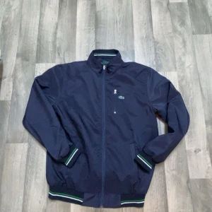 Mörkblå vindjacka från Lacoste - Snygg mörkblå vindjacka från Lacoste med klassisk krokodillogga på bröstet. Jackan har dragkedja framtill, ribbade muddar med gröna och vita ränder samt hög krage med broderad Lacoste-text. Perfekt för dig som gillar sportig och stilren look.