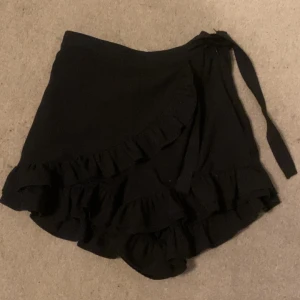 Svart omlottkjol med volang från Bik Bok - Supersöt svart kort omlottkjol från Bik Bok med volangdetaljer längs kanten och knytband i midjan, inbyggda shorts. Kjolen har en flowy känsla och passar perfekt till sneakers eller boots. Enkel att styla och ger en cool vibe till din outfit.
