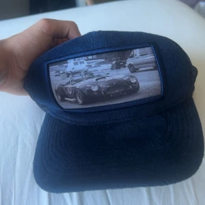 Blå keps Emilio Pendenza med bilmotiv - Snygg marinblå keps från Emilio Pendenza med mesh på baksidan och ett coolt svartvitt biltryck framtill. Kepsen har böjd skärm och justerbar passform. Perfekt för dig som gillar bilar och streetstyle. Priset kan diskuteras vid snabb affär
