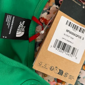 Grön cropad linnetopp North Face - Snygg grön cropad linnetopp från The North Face med smala axelband och vit logga framtill. Modellen är enkel och sportig, perfekt till jeans eller shorts. Tillverkad i mjuk bomull som känns skön mot huden.