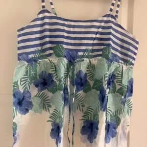 Randig och blommig sommarklänning KappAhl - Superfin kort sommarklänning från KappAhl med blåvita ränder upptill och stora blå blommor med gröna blad nedtill. Justerbara axelband och knyt i midjan. Perfekt för varma dagar och har en luftig passform i bomull.