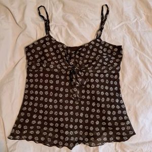 Brun blommig meshtopp med knyt - Supersöt brun meshtopp med vita blommor och smala axelband. Toppen har omlottkänsla vid bysten och en snygg knytning framtill. Materialet utanpå är i mesh men det finns även ett undre lager som är i en mer potent färg. Om ni vill se fler bilder eller har en fråga är det bara att höra av sig <3