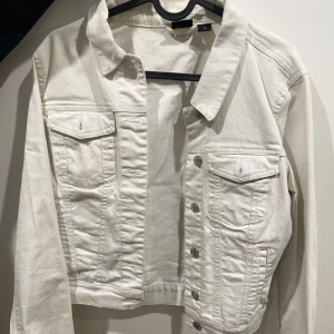 Vit jeansjacka med knappar - Snygg vit jeansjacka med klassisk krage och silverfärgade knappar framtill. Jackan har två bröstfickor med lock och knapp samt raka sömmar för en stilren look. Perfekt att slänga över en hoodie eller t-shirt.