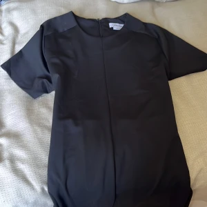 Svart långklänning från Calvin Klein - Stilren svart långklänning från Calvin Klein med korta ärmar och rund halsringning. Klänningen har en rak, elegant siluett och dragkedja i ryggen. Perfekt för dig som gillar minimalistisk och klassisk design.