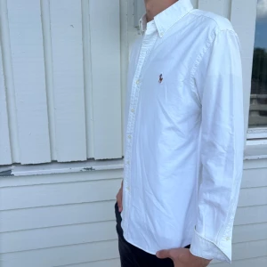 Ralph Lauren skjorta  - Klassisk vit skjorta från Ralph Lauren i modellen Custom Fit. Skjortan har button down-krage, broderad logga på bröstet och stängs med vita knappar. Tillverkad i bomull och har lång ärm. Perfekt för en clean och stilren look.