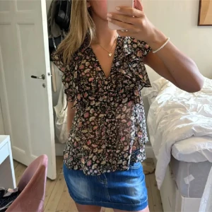 Blommig volangblus med v-ringning - Supersöt blommig blus i svart med små rosa, vita och gröna blommor. Blusen har v-ringning, volangdetaljer längs axlar och bröst samt ärmlös design. Tunt och luftigt material, perfekt för varma dagar. Snygg att styla med kjol eller jeans. Säljer både toppen och kjolen💓