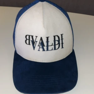 Blå och vit keps från BVALDI - Snygg truckerkeps från BVALDI med vit front och blått broderat BVALDI-tryck. Skärmen och mesh-panelerna är marinblå, vilket ger en fräsch kontrast. Justerbar snapback baktill och coolt BVALDI-band på insidan. Perfekt accessoar för en streetwear-look.