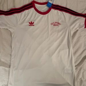 Vit CR Flamengo Adidas fotbollströja - Snygg vit fotbollströja från Adidas med CR Flamengo Brasil-brodyr på bröstet. Tröjan har röda detaljer, som krage, ärmslut och de klassiska Adidas-ränderna på axlarna. Tillverkad i lätt och bekvämt material, perfekt för fotboll eller streetwear.