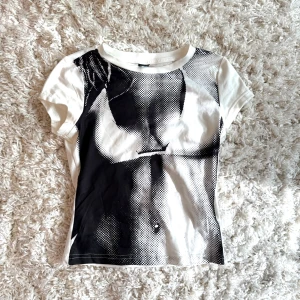 Vit och svart croppad t-shirt med tryck - Säljer en vit croppad t-shirt med svart grafiskt tryck framtill. T-shirten har korta ärmar och tight passform. Perfekt för dig som gillar unika och edgy prints.