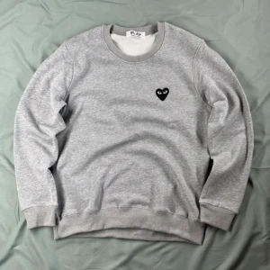 Grå sweatshirt från Comme des Garçons PLAY - Säljer en stilren grå sweatshirt från Comme des Garçons PLAY med det ikoniska svarta hjärta-loggan broderad på bröstet. Tröjan är i 100% ull och har rund halsringning samt ribbade muddar vid ärmslut och nederkant. Det är M passar S. Från nk