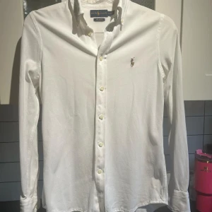 Vit långärmad skjorta från Ralph Lauren - Säljer en klassisk vit långärmad skjorta från Ralph Lauren. Pris går att prata om. Skjortan har en broderad logga på bröstet och knappar 