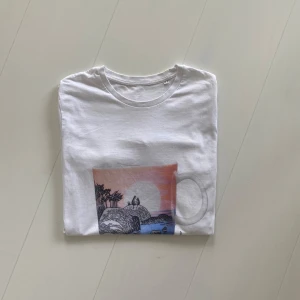 Vit t-shirt, Mumindagen 2018 - Vit t-shirt med ett färgglatt tryck framtill som föreställer en solnedgång över havet och natur. På ryggen står det '9.8.2018 by ARABIA' tryckt i svart. Klassisk rund hals och korta ärmar. Samlar objekt!