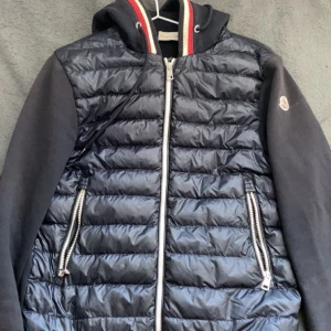 Svart pufferjacka från Moncler med huva - Säljer en svart pufferjacka från Moncler med huva och dragkedja. Jackan har quiltad framsida i glansigt material, ärmar i tyg och två fickor med dragkedja. Klassisk Moncler-logga på ärmen och sportiga detaljer vid kragen.