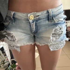 Ljusa jeansshorts med paljettdetaljer - Säljer ett par ljusblå jeansshorts med slitna detaljer och glittriga paljetter under de trasiga partierna framtill. Shortsen har fransig kant och klassisk femficksmodell. Perfekta för sommaren!
