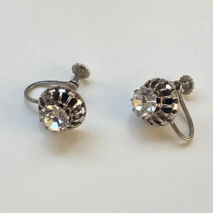 Vintage silverfärgade örhängen med skruv - Säljer ett par klassiska silverfärgade örhängen med gnistrande stenar i mitten. Örhängena har skruvstängning och en vacker korgformad infattning som ger en retro känsla. Perfekta för dig som gillar tidlös stil och elegans.