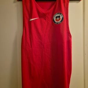 Röd Chile Linne  från Nike - Säljer en röd ärmlös Linne  från Nike med Chile-emblem på bröstet. Tröjan är tillverkad i Dri-Fit material som andas och har vita Nike-loggor både fram och bak. Perfekt för träning eller match.