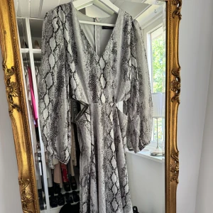 Långklänning med ormmönster och cut-outs - Säljer en långklänning med svart och grått ormmönster. Klänningen har v-ringning, långa puffärmar och snygga cut-outs i midjan. Perfekt för dig som vill sticka ut med en trendig look! klänningen är enbart använd 1 gång. 