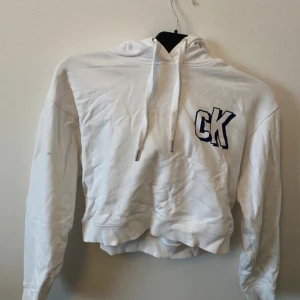Vit croppad hoodie från Calvin Klein Jeans - Säljer en vit croppad hoodie från Calvin Klein Jeans med CK-logga på bröstet och text på ena ärmen. Tröjan har huva med dragsko och ribbade muddar. Perfekt för en avslappnad och trendig look.Frakt tillkommer