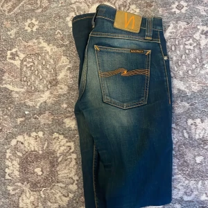 Mörkblå jeans från Nudie Jeans - Säljer ett par mörkblå jeans från Nudie Jeans med klassiska orange sömmar och femficksmodell. Byxorna har en smal passform och raka ben. Perfekta för dig som gillar stilrena och tidlösa jeans.
