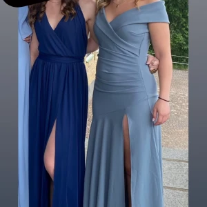 Ljusblå offshoulder långklänning med slits - Säljer en elegant ljusblå långklänning med offshoulder-skärning och draperad överdel. Klänningen har en hög slits framtill och figurnära passform som framhäver siluetten. Perfekt för bal eller festliga tillfällen.