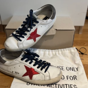 Golden Goose Superstars - Säljer ett par vita Golden Goose sneakers med en glittrig röd stjärna på sidan, mörkblå snörning och grå hälkappa. Skorna är köpta i Grekland år 2023 från affärerna Soho-Soho.  Box, klistermärken, påse och kvitto ingår!👊 Köpta för €460 vilket motsvarar 5000 Kr. Skick 9/10, inte använt så ofta. Frakt står köparen för.