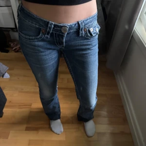 True Religion lågmidjade bootcut jeans - Säljer ett par klassiska blå jeans från True Religion med låg midja och bootcut. Jeansen har tydliga kontrastsömmar, stora fickor med lock och silverfärgade knappar. Snygg tvätt och detaljerad brodyr på bakfickorna samt logga på bakre linningen. Jag säljer dessa eftersom de är lite för korta på mig som är 167 cm, de är omsydda så de är lite utsvängda !