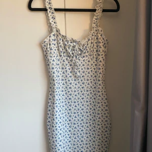 Vit blommig klänning från SHEIN - Söt vit klänning från SHEIN med små blå blommor och volangdetaljer på axelbanden. Klänningen har knytning framtill och är ärmlös med en smickrande passform. Perfekt för varma dagar och somriga tillfällen.