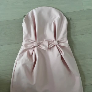 Ljusrosa axelbandslös klänning  - Söt axelbandslös klänning i ljusrosa med två dekorativa rosetter framtill i midjan. Köpt för två veckor sen och endast använd en gång, nypris 785kr