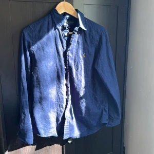 Mörkblå linneskjorta från Ralph Lauren - Säljer en mörkblå skjorta i linne från Ralph Lauren med klassisk krage och knappar framtill. Skjortan har lång ärm och den ikoniska broderade loggan på bröstet. Perfekt för en stilren och avslappnad look. En knapp saknas på den högra ärmen men det finns extra knappar. Så det är en lätt lagning 