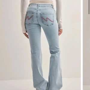 Ljusblå bootcut jeans med slitningar - Snygga virala  ljusblå jeans med bootcut passform och slitna detaljer vid knäna. Bakfickorna har en cool broderad zigzag-detalj i rosa. Klassisk femficksmodell med knapp och dragkedja framtill. (Helt nya å ligger i förpackningen med prislapp på 