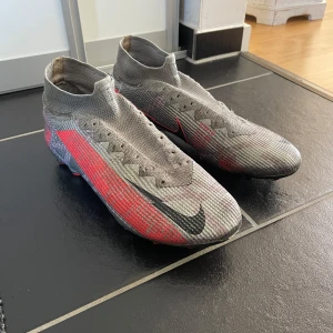 Gråa Nike fotbollsskor super fly 7 Elite - Säljer ett par gråa Nike fotbollsskor med högt skaft och röda samt svarta detaljer. Skorna plös med integrerad strumpa för extra stöd. Sulan har fasta dobbar för spel på gräs. Använda men okej skick, snören ingår inte.