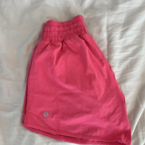 Rosa träningsshorts från Lululemon - Säljer ett par knallrosa träningsshorts från Lululemon med bred resår i midjan och ventilerande meshdetaljer på sidorna. Perfekta för löpning eller gym. Klassisk Lululemon-logga på benet. Osäker på storlek men tror 2, sitter som xs-s