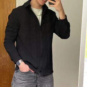 Svart overshirt med dragkedja - Säljer en stilren svart overshirt med dragkedja framtill och klassisk krage. Jackan har två bröstfickor och långa ärmar. Perfekt att slänga över en t-shirt för en clean look.