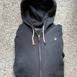 Svart hoodie med dragkedja från Morris - Svart hoodie från Morris med dragkedja framtill och justerbar huva med snören. Klassisk design med liten broderad logga på bröstet. Perfekt för en avslappnad stil.