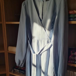 Ljusblå omlottklänning med knytband - Säljer en ljusblå kortklänning med omlottmodell och knytband i midjan. Klänningen har långa ärmar och v-ringad halsringning. Perfekt för vår och sommar, enkel men stilren design. Använd 1 gång, nypris 169kr